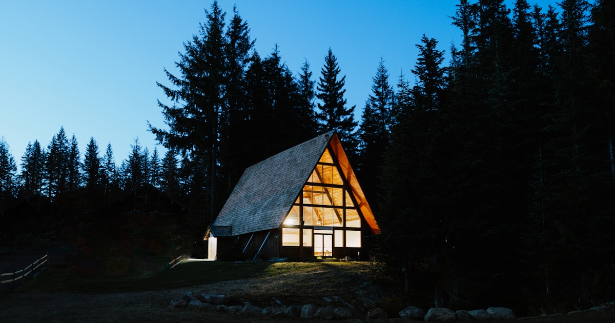 An A-frame cabin in the forest at dusk with the lights on — everything already built